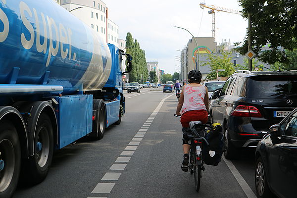 Das Bild zeigt eine Radfahrerin von hinten auf einem Radweg. Rechts von ihr eine Reihe parkender Autos, links von ihr fährt ein LKW dicht an ihr vorbei.