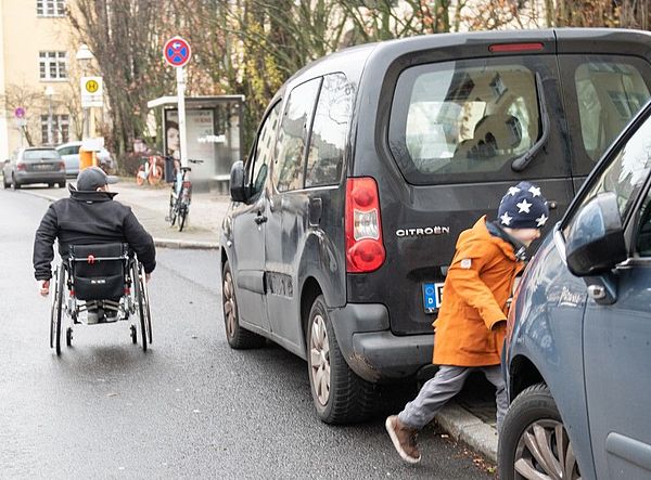Das Bild zeigt zwei Autos, die aufgesetzt auf einem Gehweg parken. Ein kleiner Junge geht zwischen den Autos hindurch, ein Rollstuhlfahrer fährt auf der Straße an den Autos vorbei.